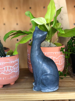 A concrete cat sculpture facing away to show its back view, on a shelf with plants. 